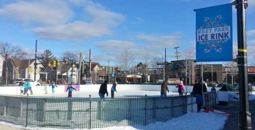 A winter scene at Riley Park Ice Rink with people skating on ice under a blue sky and a sign for the rink.