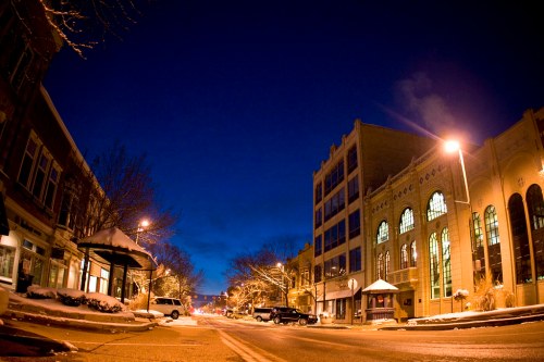 A snowy downtown street at dusk, with buildings lining the road and streetlights illuminating the scene.