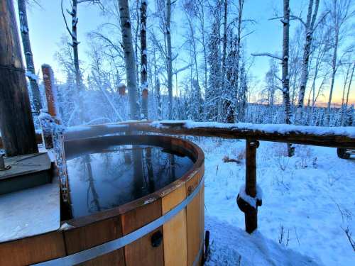 A wooden hot tub surrounded by snow-covered trees, with steam rising against a serene winter landscape.