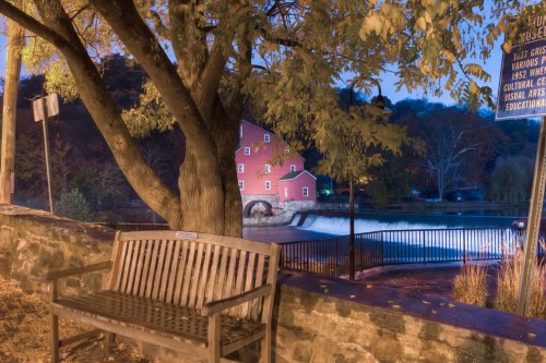 A wooden bench beside a stone wall, with a red mill and water feature in the background, surrounded by autumn foliage.