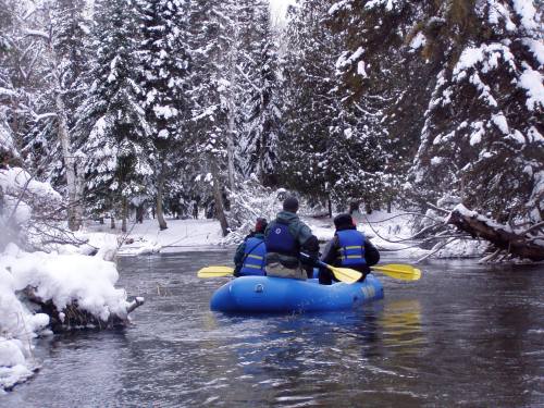 A group of people in a blue raft paddling through a snowy forested area along a river.