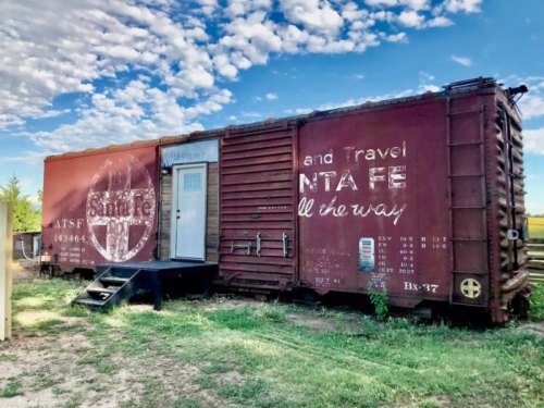 A vintage red train car converted into a small travel accommodation, set against a blue sky with scattered clouds.