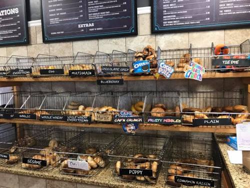 A display of various bagels in baskets, labeled with flavors like blueberry, chocolate chip, and cinnamon raisin.