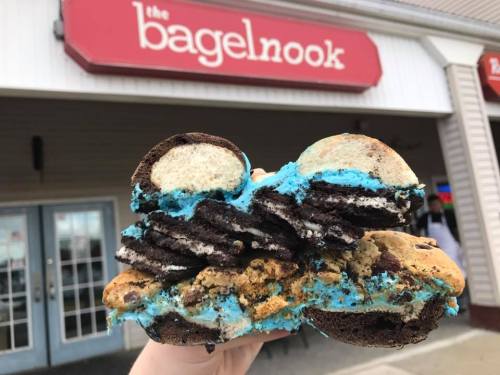 A hand holds a large, colorful dessert sandwich made of cookies and blue frosting, in front of a bagel shop.