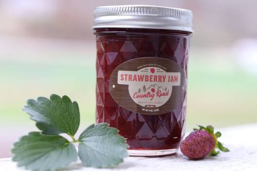 A jar of strawberry jam with a diamond-patterned design, labeled "Country Road," next to a fresh strawberry and leaves.