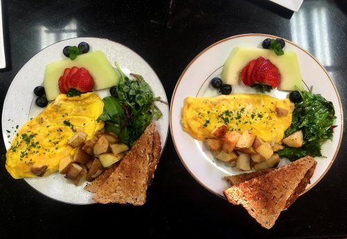 Two plates of breakfast featuring omelets, sautéed greens, roasted potatoes, toast, and fresh fruit.
