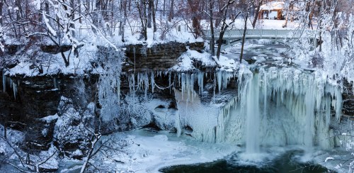 A frozen waterfall surrounded by snow-covered trees and icicles, with a bridge in the background.