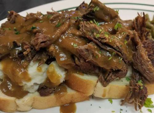 A plate of tender beef covered in gravy, served over mashed potatoes and white bread, garnished with herbs.