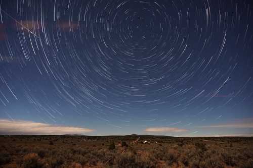 Star trails swirl in the night sky over a desert landscape, with distant mountains and sparse vegetation visible below.