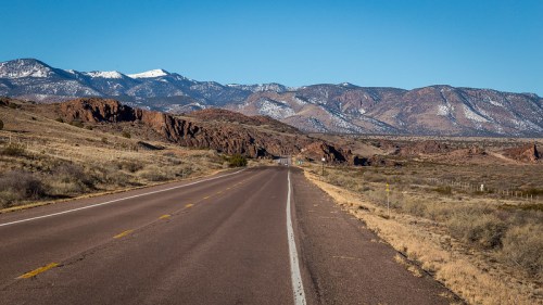 A long, empty road stretches through a desert landscape, with mountains and snow-capped peaks in the background.