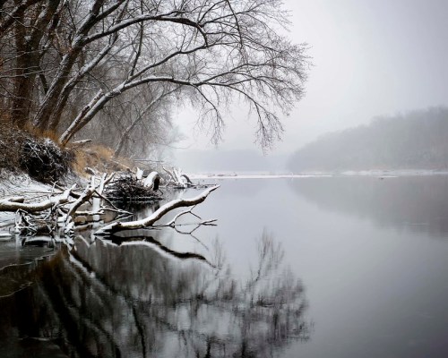 A serene winter landscape featuring a snowy riverbank, bare trees, and reflections on a calm, misty river.