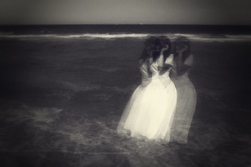 A ghostly figure in a white dress stands in the ocean, reflecting on the water's surface under a moody sky.