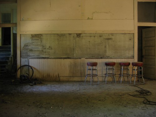 Abandoned room with four red bar stools against a weathered wall, featuring peeling paint and debris on the floor.