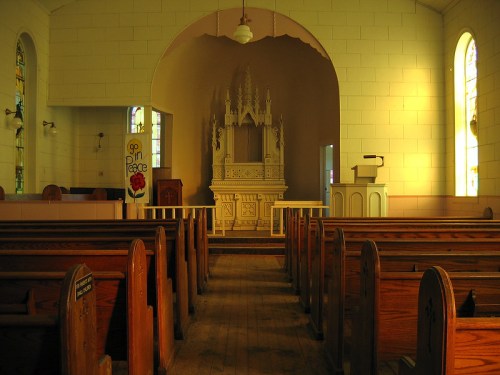 Interior of a church featuring wooden pews, a white altar, and stained glass windows, illuminated by soft light.