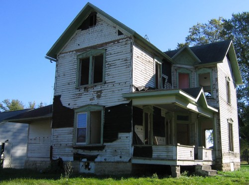 An old, weathered two-story house with peeling paint and broken windows, surrounded by overgrown grass.