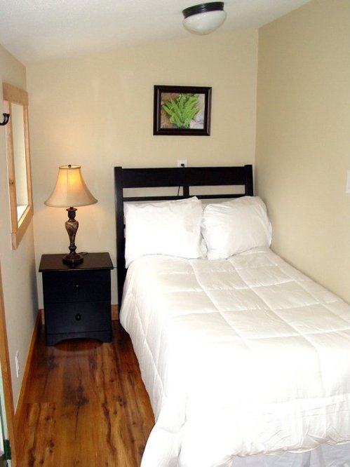 Cozy bedroom with a single bed, nightstand, lamp, and a framed plant picture on the wall. Warm wood flooring.