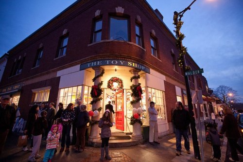 A festive toy shop at dusk, adorned with holiday decorations, with a crowd of people gathered outside.