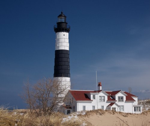 A tall black and white lighthouse beside a white house, set against a clear blue sky and sandy dunes.
