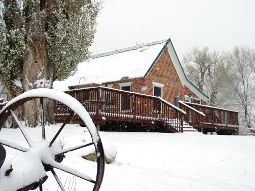 A snow-covered house with a wooden deck, surrounded by trees and a wagon wheel in the foreground.