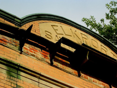 Close-up of a weathered brick building sign reading "Elkport Saloon," with greenery in the background.