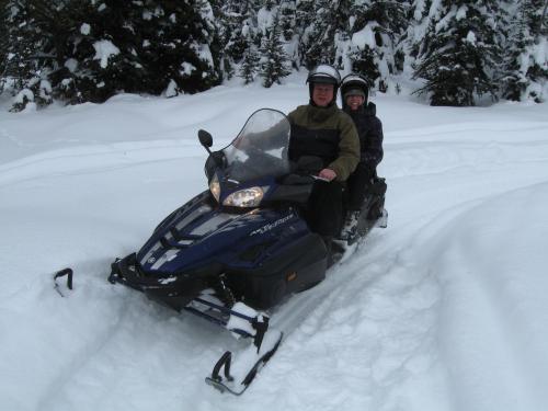 A man and a woman ride a blue snowmobile through a snowy landscape surrounded by trees.
