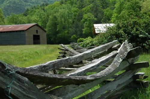 A rustic wooden fence lines a grassy path, with green trees and two barns in the background under a clear sky.