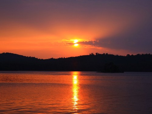 Sunset over a calm lake, with vibrant orange and purple hues reflecting on the water and silhouetted hills in the background.