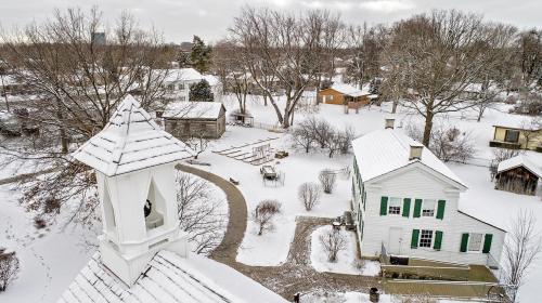 A snowy landscape featuring a white house, trees, and a winding path, with a bell tower in the foreground.
