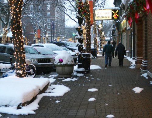 Snow-covered street with holiday lights, parked cars, and two people walking past a restaurant.
