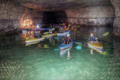 A group of kayakers with headlamps paddles through a dimly lit cave with shimmering water.