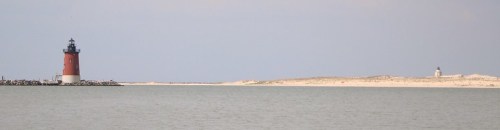 A red and white lighthouse stands on the left, with a sandy shore and a distant lighthouse on the right, under a clear sky.