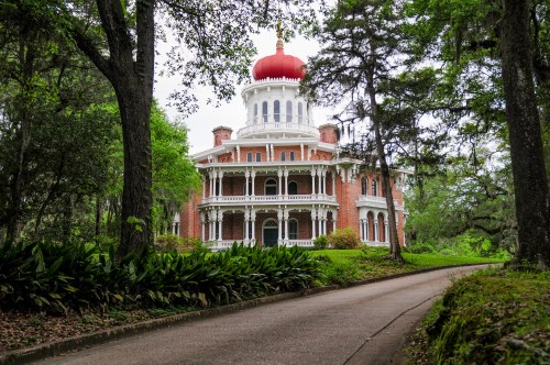 A grand, red-domed mansion surrounded by lush greenery and trees, with a winding path leading to its entrance.