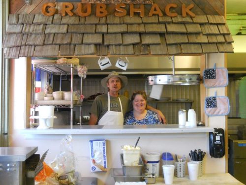 A man and woman smile together behind the counter of a cozy eatery called "Grub Shack."
