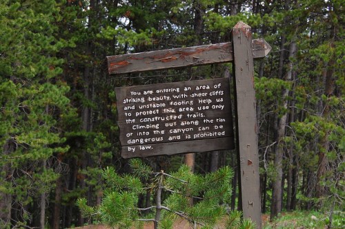 Sign warning about dangerous cliffs and unstable footing, advising to use only constructed trails in a forested area.