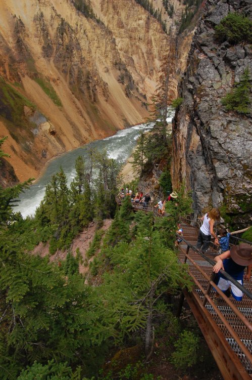 A winding wooden path along a cliffside, with people walking and a river flowing below, surrounded by trees and rocky terrain.
