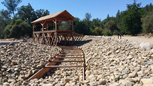 A wooden structure on a rocky landscape, surrounded by trees under a clear blue sky.