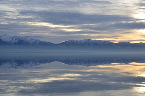 A serene landscape featuring snow-capped mountains reflected in calm water under a cloudy sky at sunset.