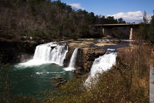 A scenic view of a waterfall cascading into a river, with a bridge in the background and trees lining the shore.