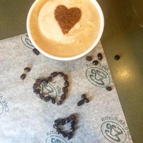 A cup of coffee with a heart-shaped foam design, surrounded by coffee beans arranged in heart shapes on a paper.