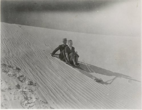 Two people sitting on a sled, sliding down a sandy dune in a vintage black-and-white photograph.