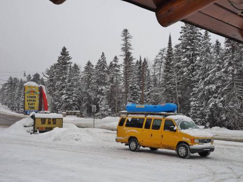 A yellow van with a blue kayak on top parked in a snowy landscape with trees and a sign for outdoor activities.