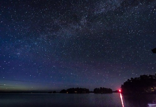 A starry night sky over a calm lake, with distant trees silhouetted and a faint red light reflecting on the water.
