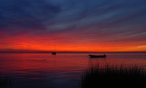A serene sunset over a calm lake, with vibrant orange and purple hues reflecting on the water and silhouettes of boats.