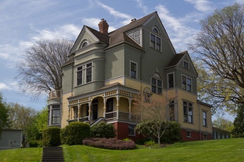 A large, colorful Victorian-style house with a wraparound porch, surrounded by lush greenery and blue skies.