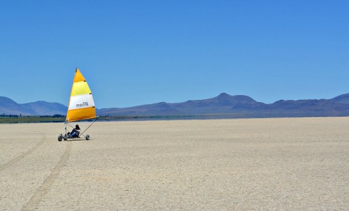 A person sails on a land yacht across a vast, dry salt flat under a clear blue sky. Mountains are visible in the background.