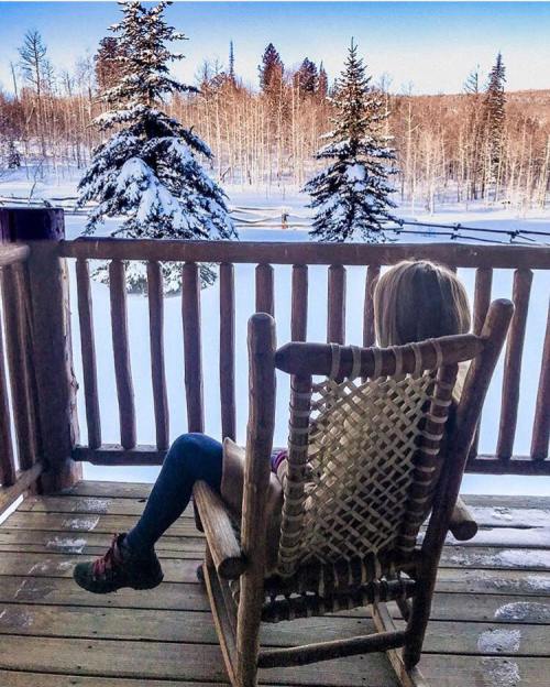 A person sits in a rocking chair on a snowy balcony, overlooking a winter landscape with trees.