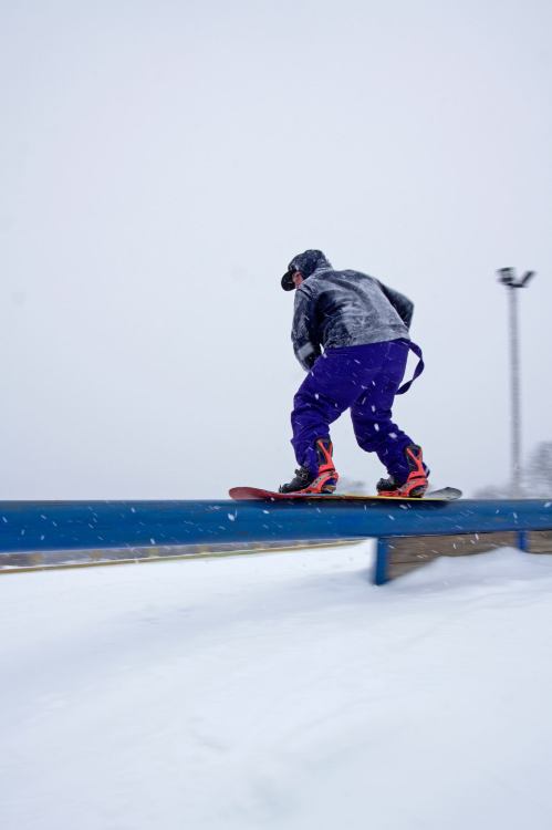 A snowboarder in purple pants rides a blue rail in snowy conditions, with snowflakes falling around.
