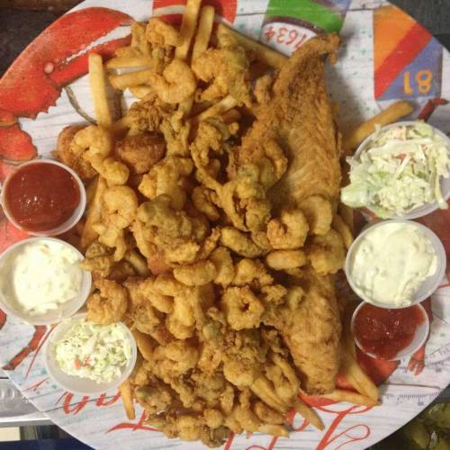 A large platter of fried seafood, including shrimp, fish, and fries, served with dipping sauces and coleslaw.