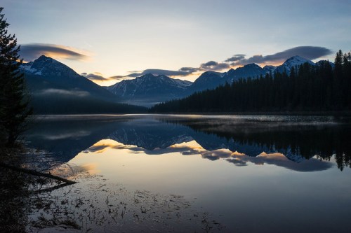 Serene lake at sunrise, reflecting mountains and clouds, surrounded by trees and mist.