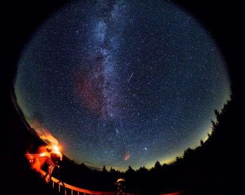 A starry night sky filled with the Milky Way and a shooting star, viewed from a dark outdoor setting.